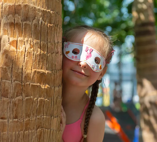 Young girl wearing a decorated paper mask peeks from behind a tree trunk, sunny playful mood.