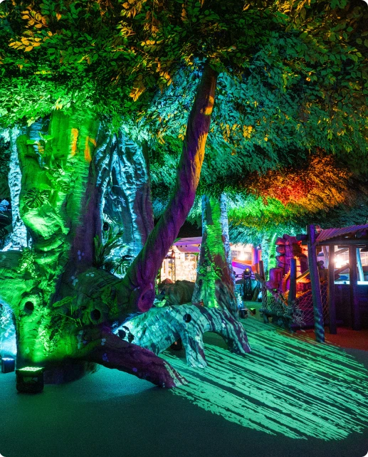 Large artificial tree lit with green and purple lights inside a whimsical indoor play area.