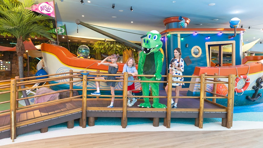 Four children and a mascot on a boardwalk in an indoor play area beside a decorative boat, smiling.