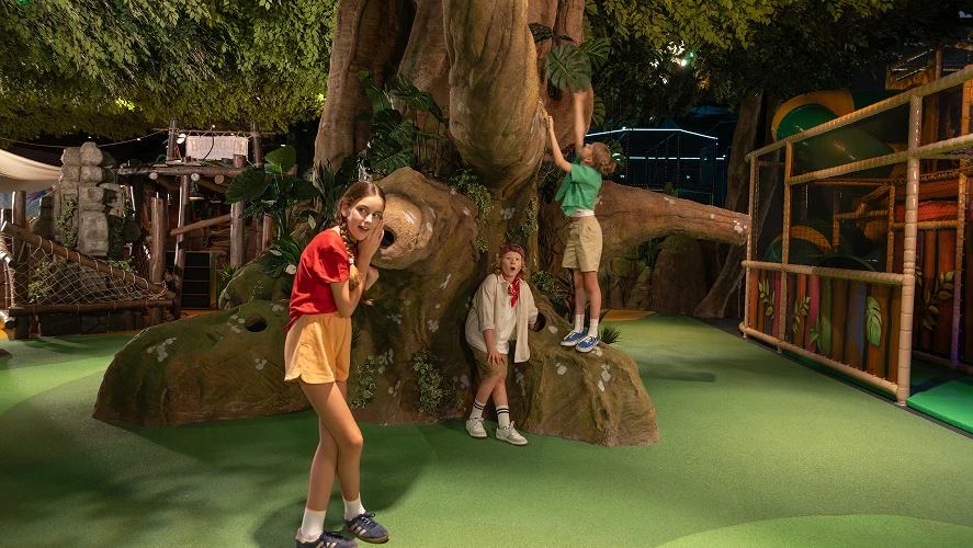 Kids climbing and exploring an oversized tree structure inside an indoor adventure playground, joyful atmosphere.