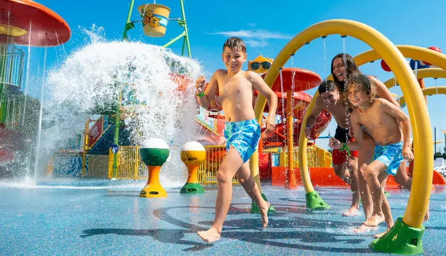 A family runs under water arches in an outdoor splash zone, with colorful slides and fountains in the background.