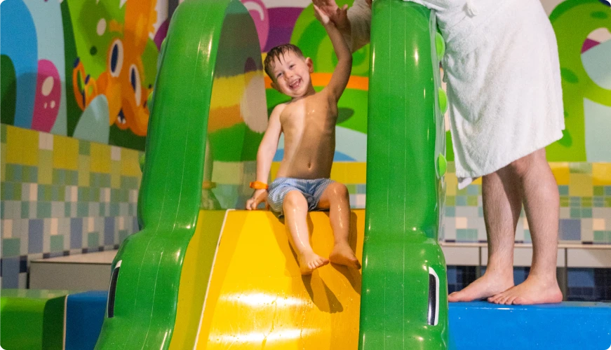 A boy sliding down a colourful slide at a water park, smiling, with a parent's assistance