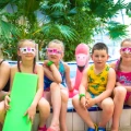 Group of smiling schoolchildren in swimming costumes and carnival masks at a swimming pool surrounded by palm trees