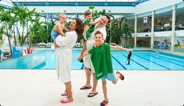 A family of five wearing bathrobes and flip-flops having fun next to an indoor swimming pool surrounded by palms